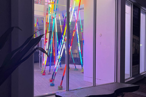 image of a light installation looking through a window. Light installation is made up of clear acrylic tubes that run from floor to ceiling with LED lights running the length of each tube. Lights are blue, red, yellow, green.