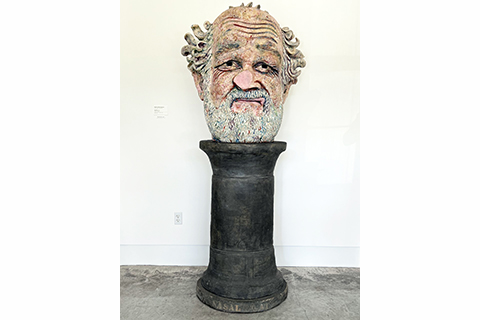 A seven-foot-tall pottery and glaze sculpture depicting a bearded head atop a simple black pedestal. The head is that of an older man with medium-light skin tone, pock marks on his cheeks, and deep wrinkles on his high forehead and at the corners of his eyes. His beard and mustache are gray, as is the wavy hair at the sides of his balding head. The awkward sneer, or grimace, on his lips, and the utterly smooth, flat tip of his squished, bulbous nose, evoke a face pressed against a window or pane of glass.