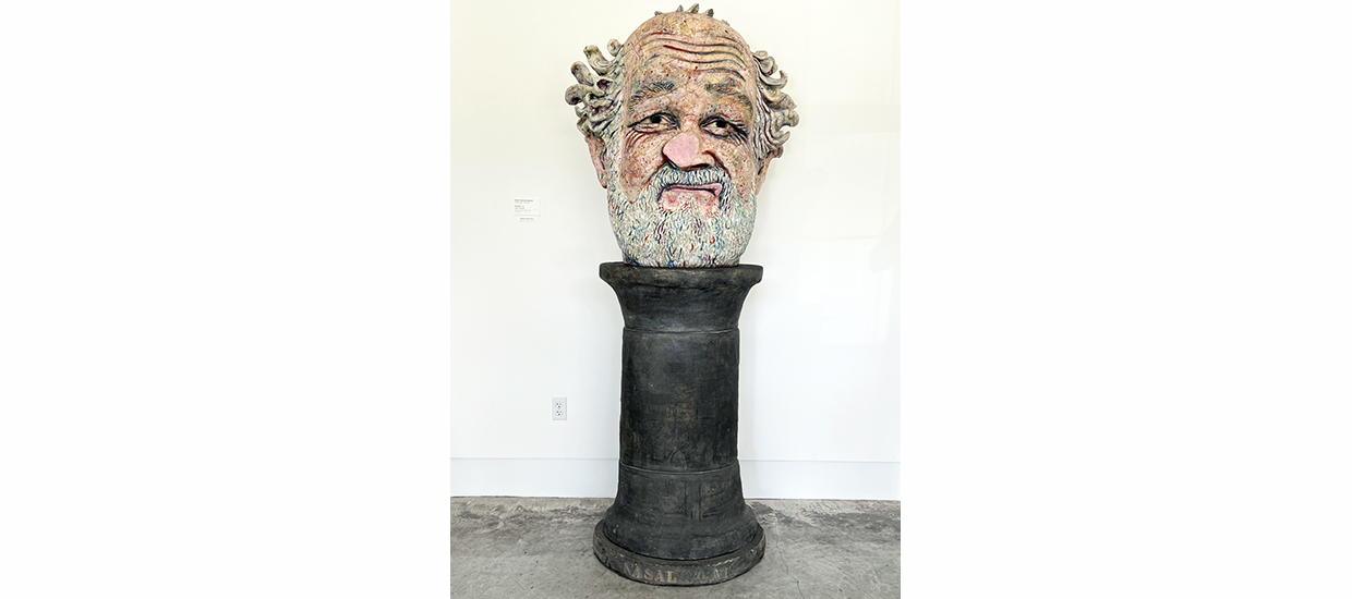 A seven-foot-tall pottery and glaze sculpture depicting a bearded head atop a simple black pedestal. The head is that of an older man with medium-light skin tone, pock marks on his cheeks, and deep wrinkles on his high forehead and at the corners of his eyes. His beard and mustache are gray, as is the wavy hair at the sides of his balding head. The awkward sneer, or grimace, on his lips, and the utterly smooth, flat tip of his squished, bulbous nose, evoke a face pressed against a window or pane of glass.