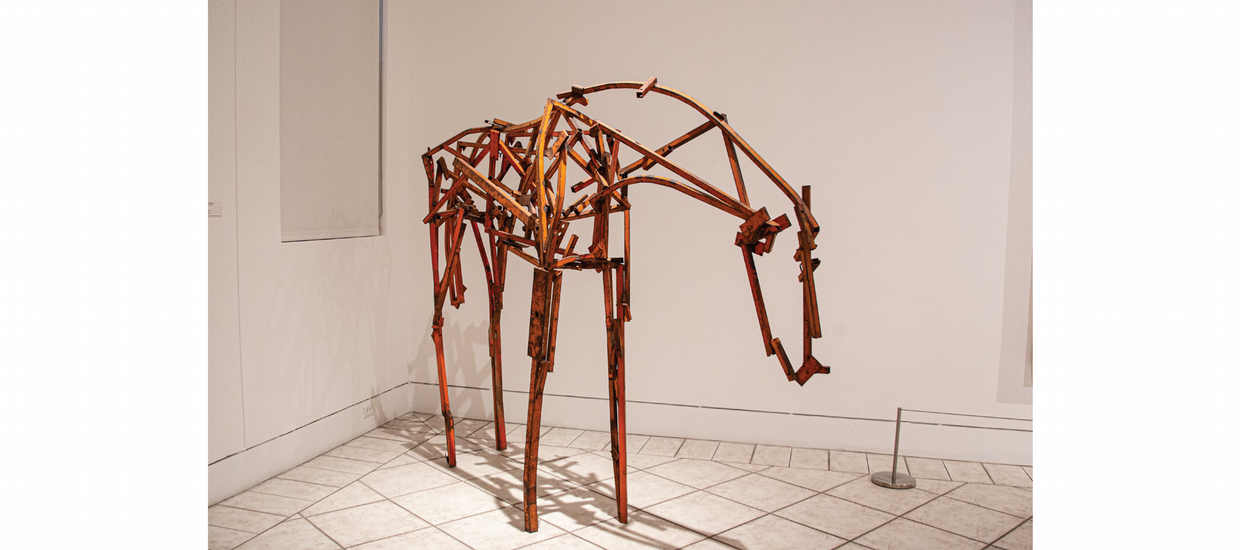 A life-sized sculpture of a horse, minimally rendered with welded strips of found steel. The horse stands on thin legs, front hooves and back hooves parallel, head down, tail tucked under the rump. The strips of curved steel creating the animal’s long neck, slightly swayed back, and round rump form an outline, the literal backbone of the sculpture. In the horse’s body, the strips of rust-colored [or brown-painted] steel are straight and have been welded together at sharp, irregular angles, creating the look of a jumbled skeleton. 