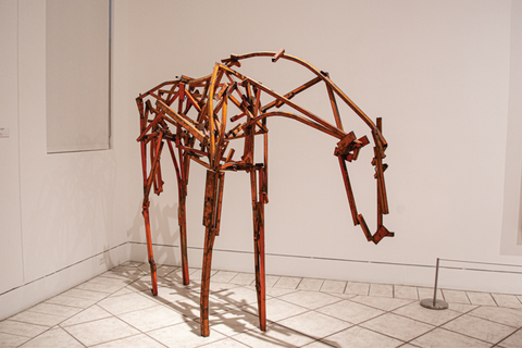A life-sized sculpture of a horse, minimally rendered with welded strips of found steel. The horse stands on thin legs, front hooves and back hooves parallel, head down, tail tucked under the rump. The strips of curved steel creating the animal’s long neck, slightly swayed back, and round rump form an outline, the literal backbone of the sculpture. In the horse’s body, the strips of rust-colored [or brown-painted] steel are straight and have been welded together at sharp, irregular angles, creating the look of a jumbled skeleton. 