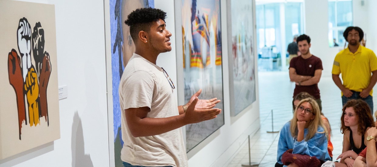 student talking to seated students in the middle of an art gallery