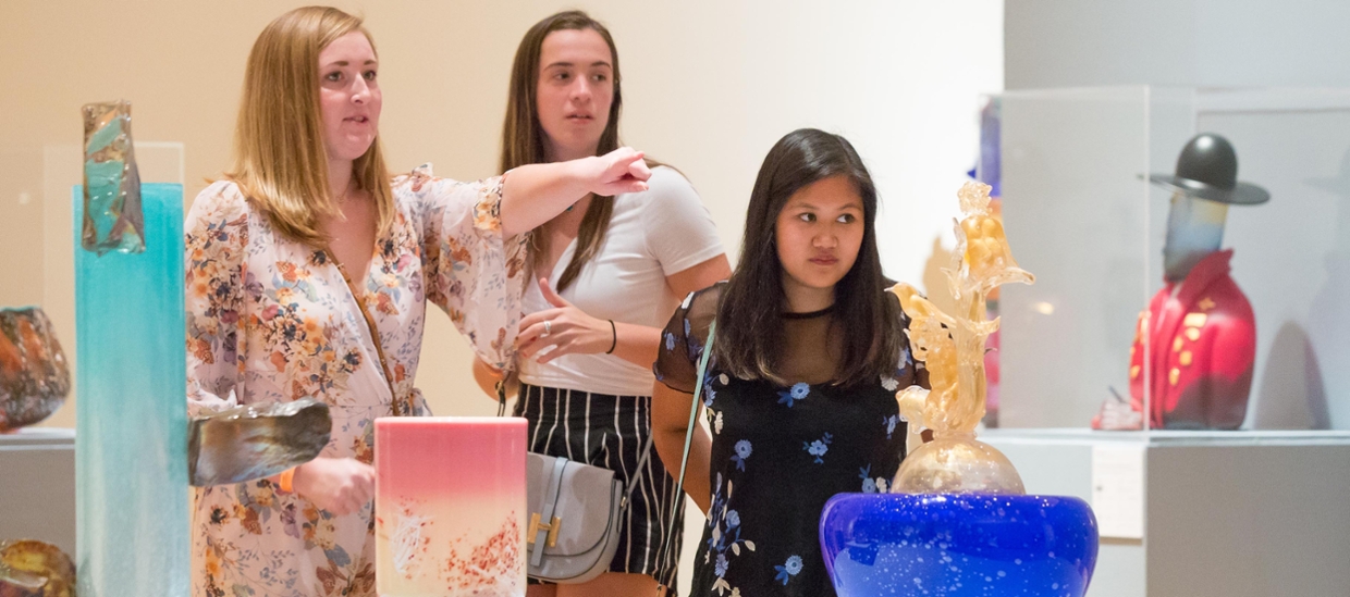 three woman looking at glass art in an art gallery. Glass objects are vase like