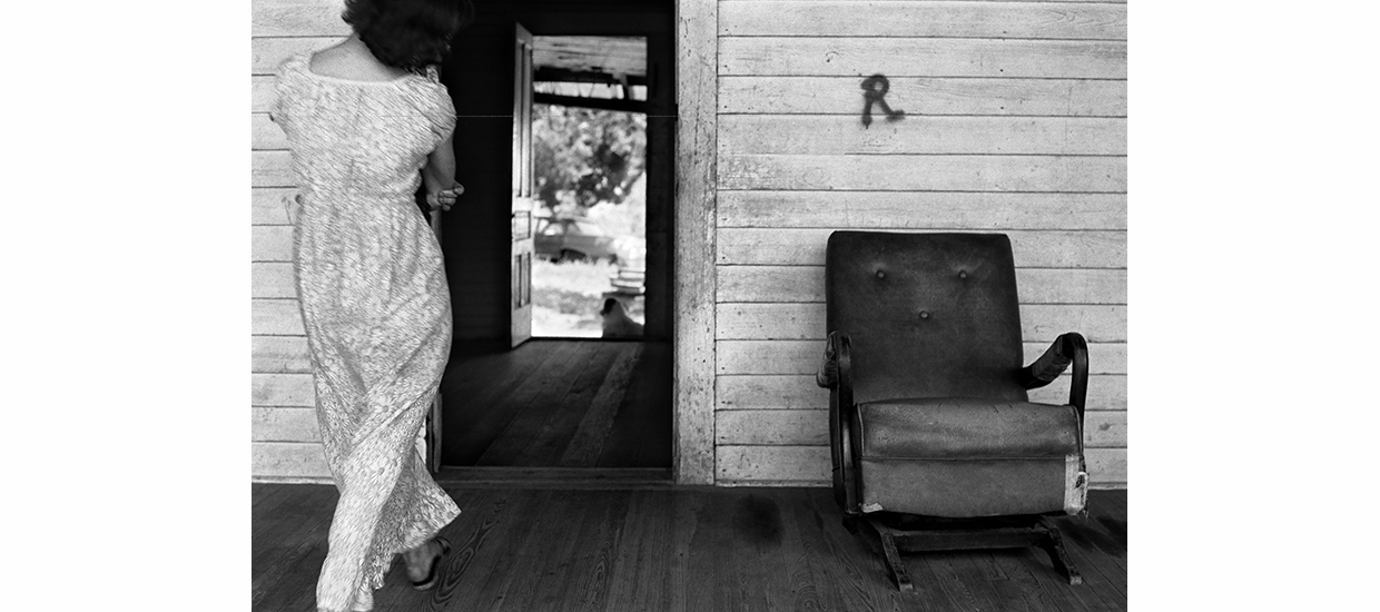 black and white photograph of a woman seen from the back walking into a doorway. There is a chair to the right