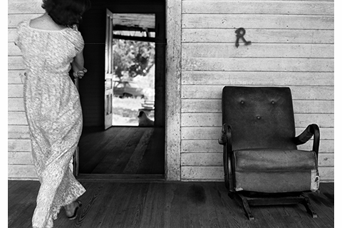 black and white photograph of a woman seen from the back walking into a doorway. There is a chair to the right