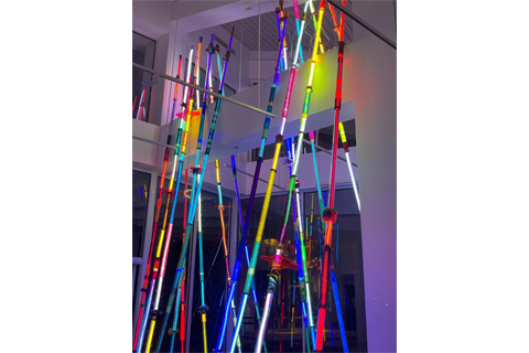 image of an art installation of clear acrylic tubes wrapped in colored fabric with LED lights illuminating the tubes. The tubes run from floor to ceiling averaging around 15 feet in height. 