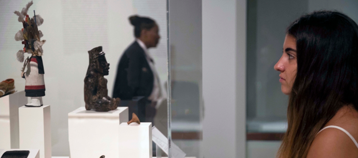 image of a woman looking into a glass case with native american art depicting small figures made of wood and cloth