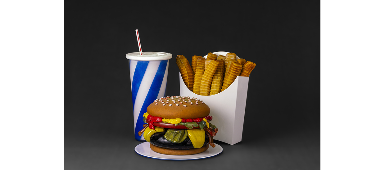 glass sculptures of realistic looking hamburger with ketchup, pickles, lettuce, a box of french fries, and a drink with a straw. The box for french fries is white and cup with straw has blue and white stripes