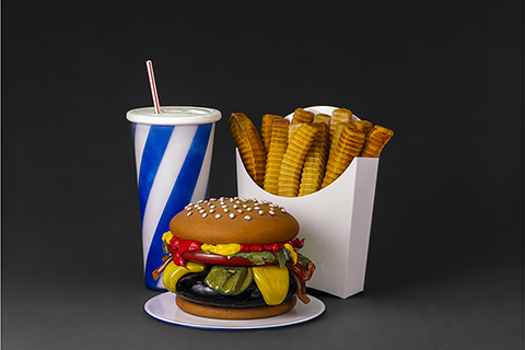 glass sculptures of realistic looking hamburger with ketchup, pickles, lettuce, a box of french fries, and a drink with a straw. The box for french fries is white and cup with straw has blue and white stripes