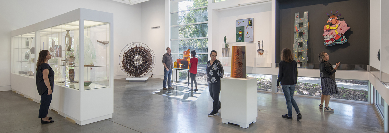 figures in a gallery with cases of contemporary glass works. Large window in the background.