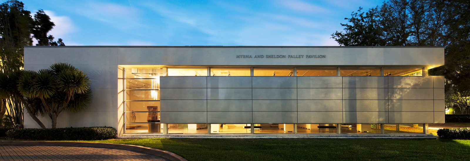 exteriro image of the palley pavillion at the Lowe Art Museum. It is dusk and the lights of the interior of the museum can be seen through windows. 