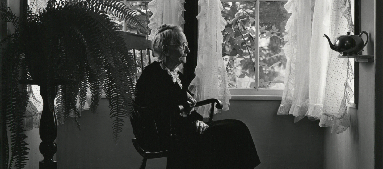 black and white photo of an elderly woman seated in a chair, profile in a room with a fern and lace curtains