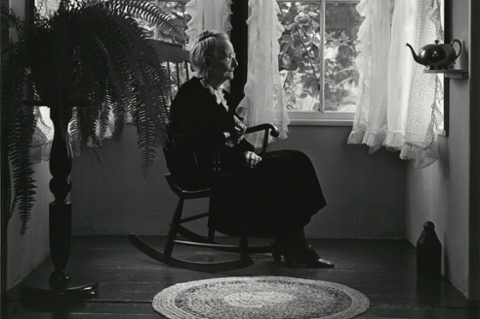 black and white photo of an elderly woman seated in a chair, profile in a room with a fern and lace curtains