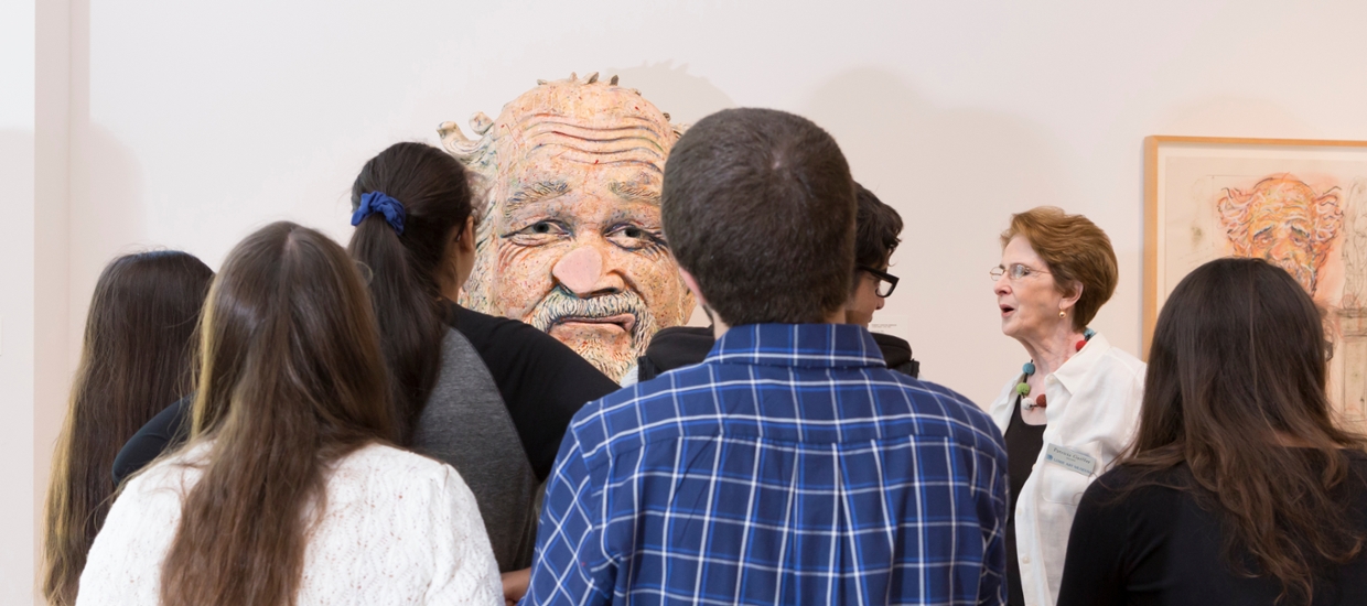 docent giving a talk in front of a group or people looking at a larger than life head made of ceramic clay. The nose of the figure is flattened as if it walked into a glass wall