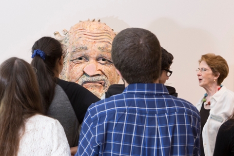 docent giving a talk in front of a group or people looking at a larger than life head made of ceramic clay. The nose of the figure is flattened as if it walked into a glass wall