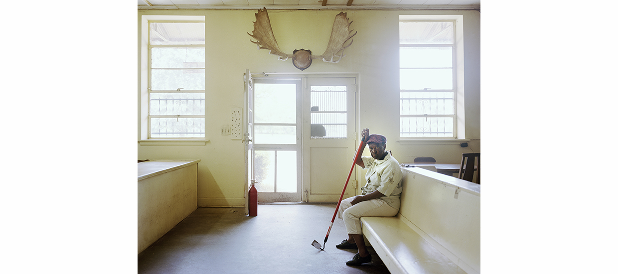 Photograph of a room in a building with white walls and what appear to be elk horns mounted on a wall above a central doorway. There is a black woman seated on a bench leanining on a long rod that looks to be part of some tool.