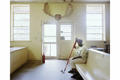 Photograph of a room in a building with white walls and what appear to be elk horns mounted on a wall above a central doorway. There is a black woman seated on a bench leanining on a long rod that looks to be part of some tool.