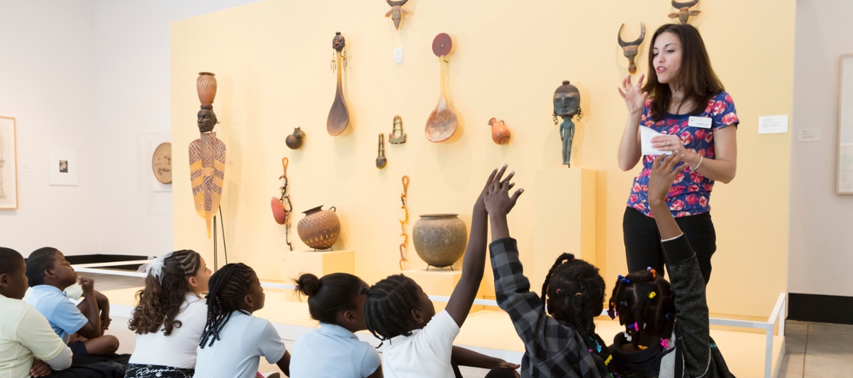 image of students looking at glass sculptures on a wall being talked to by an educator. Some students are raising their hands