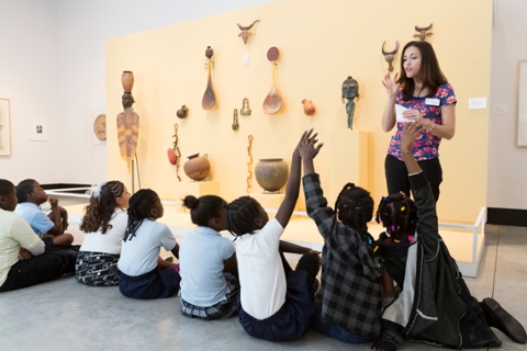 image of students looking at glass sculptures on a wall being talked to by an educator. Some students are raising their hands