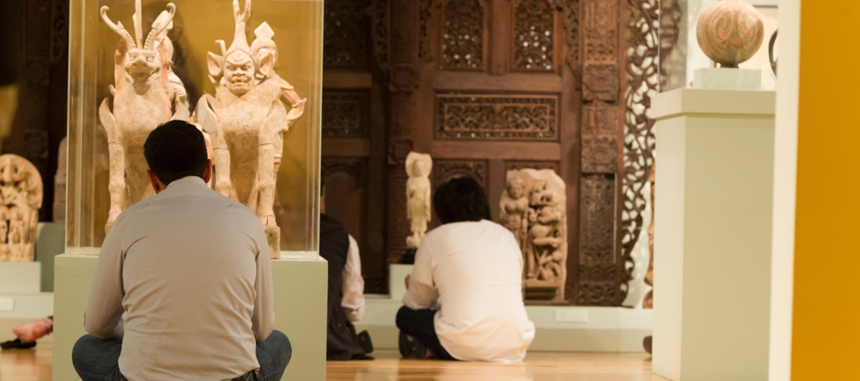 student sitting on floor of museum in Asian galleries looking at sculptures of anamorphic deities that are part dog and part human made of stone