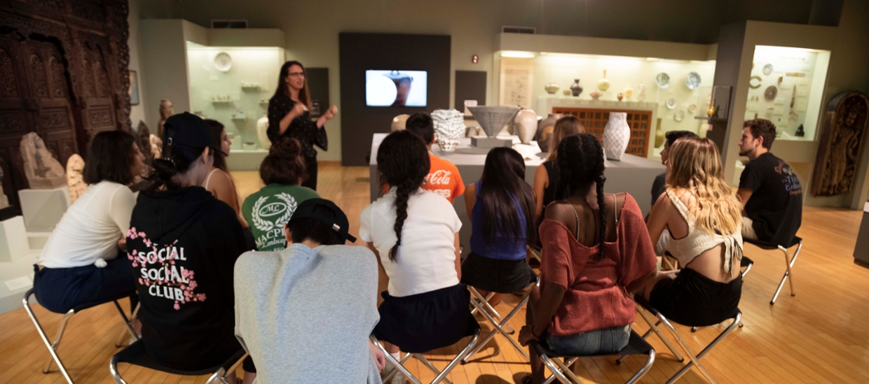 students seated in a gallery with arts from asia being spoken to by a musem guide