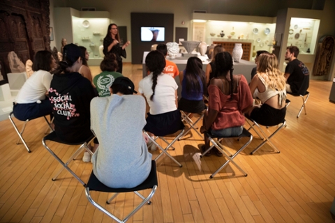 students seated in a gallery with arts from asia being spoken to by a museum guide