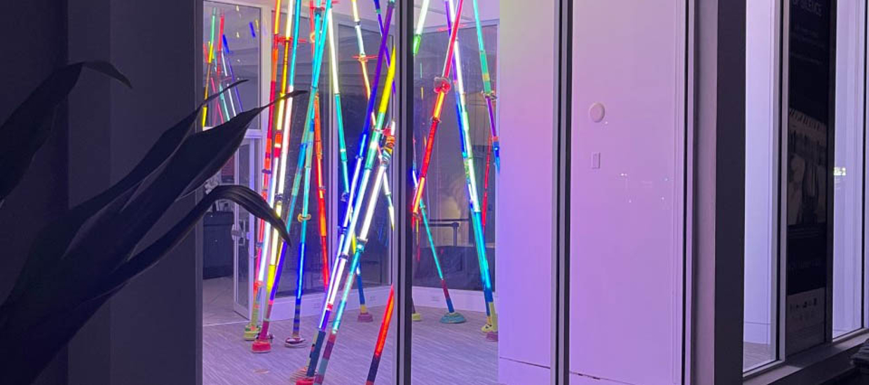 image of a light installation looking through a window. Light installation is made up of clear acrylic tubes that run from floor to ceiling with LED lights running the length of each tube. Lights are blue, red, yellow, green.